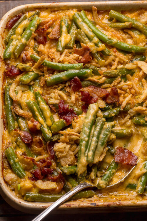 Baking dish with green bean casserole covered in a cheesy bacon sauce with a serving spoon.