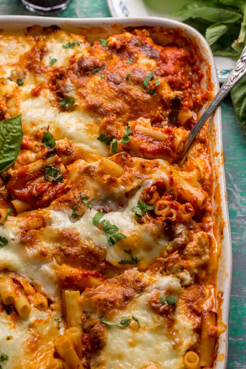 Baking dish with baked ziti in it on a table with basil and wine.